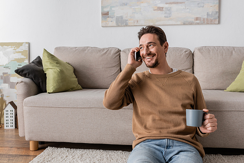 cheerful man in beige jumper talking on smartphone while sitting with cup of coffee at home