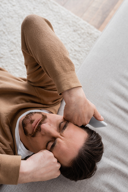 top view of tired man in beige jumper holding mobile phone and leaning on sofa
