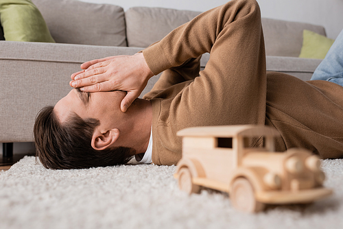 man covering face with hands and lying on carpet near wooden toy vehicle in living room