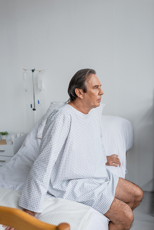 Grey haired patient in gown sitting on bed in hospital