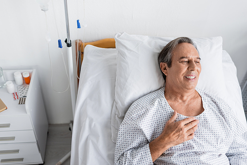 High angle view of smiling elderly man in patient gown lying on bed in hospital ward