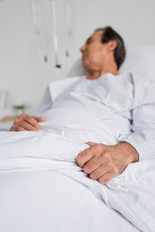 Blurred senior patient touching blanket while lying on bed in hospital