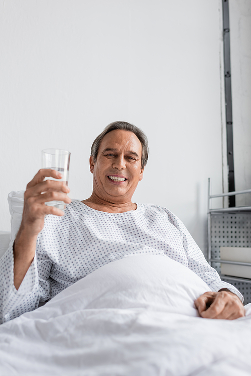 Smiling man holding glass of water and looking at camera on hospital bed
