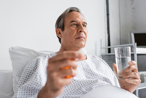 Senior man in gown holding pills and water in hospital ward