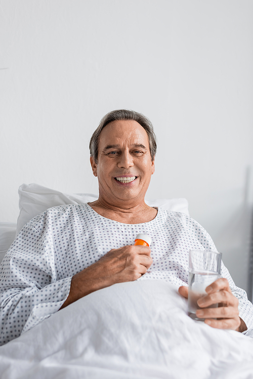 Cheerful grey haired man holding pills and water while looking at camera on hospital bed