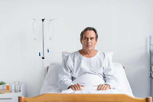 Senior patient looking at camera while sitting on bed in hospital ward