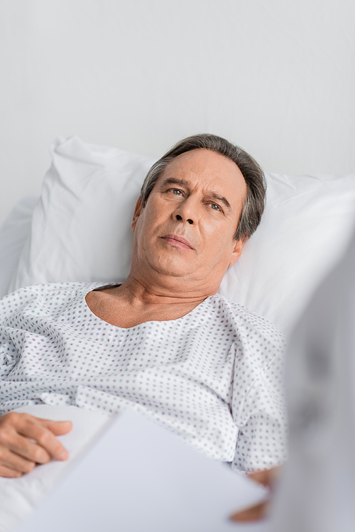 Elderly man in patient gown looking at blurred doctor in clinic