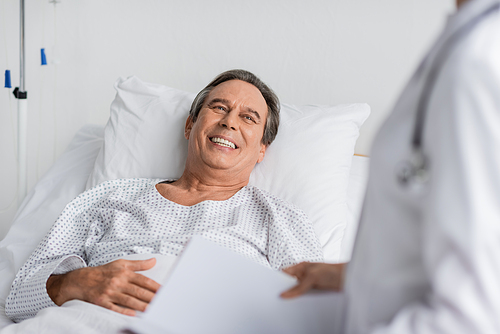 Cheerful patient in gown looking at blurred doctor with paper folder in hospital ward