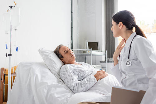 Doctor with paper folder touching neck while talking to senior patient in hospital ward