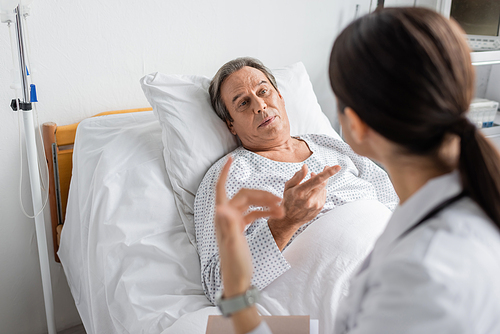 Senior patient talking to blurred doctor with paper folder in hospital ward