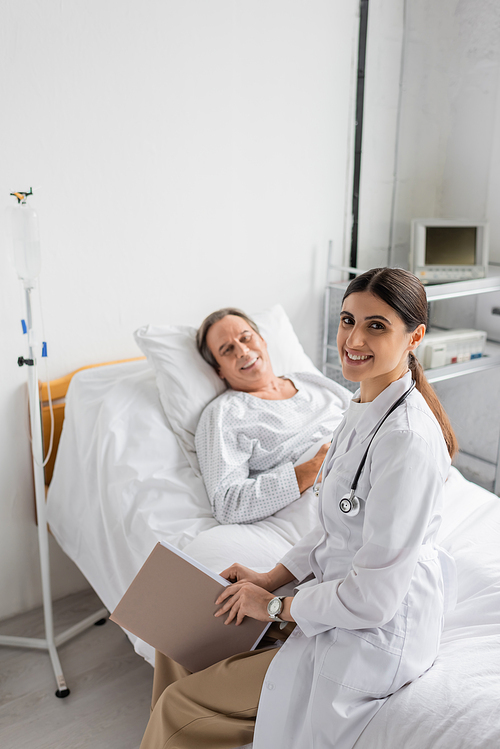 Positive doctor holding paper folder and looking at camera near blurred senior patient in clinic