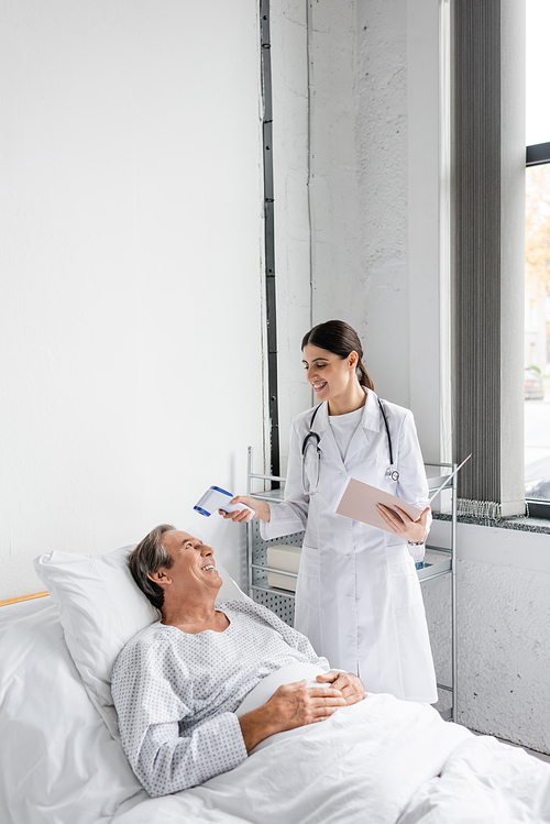 Cheerful doctor with paper folder holding pyrometer near senior patient in hospital ward