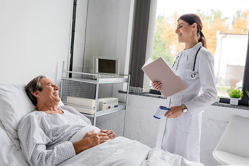 Positive elderly patient looking at doctor with paper folder and pyrometer in hospital ward