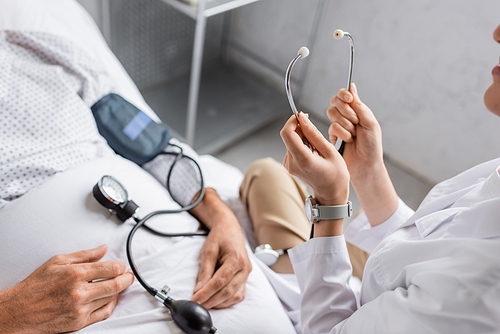 Cropped view of doctor holding stethoscope near tonometer and sick patient on hospital bed