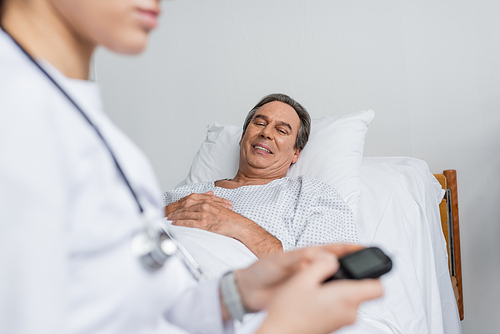 Smiling elderly patient looking at blurred doctor with glucometer in hospital ward
