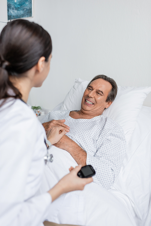Smiling senior patient looking at blurred doctor with glucometer in clinic