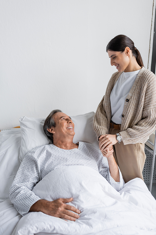 Smiling daughter holding hand of father lying on bed in hospital