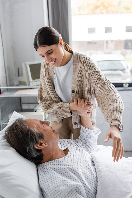 Positive woman holding hand of elderly father in patient gown in clinic