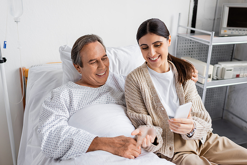 Smiling woman holding smartphone near senior father on hospital bed