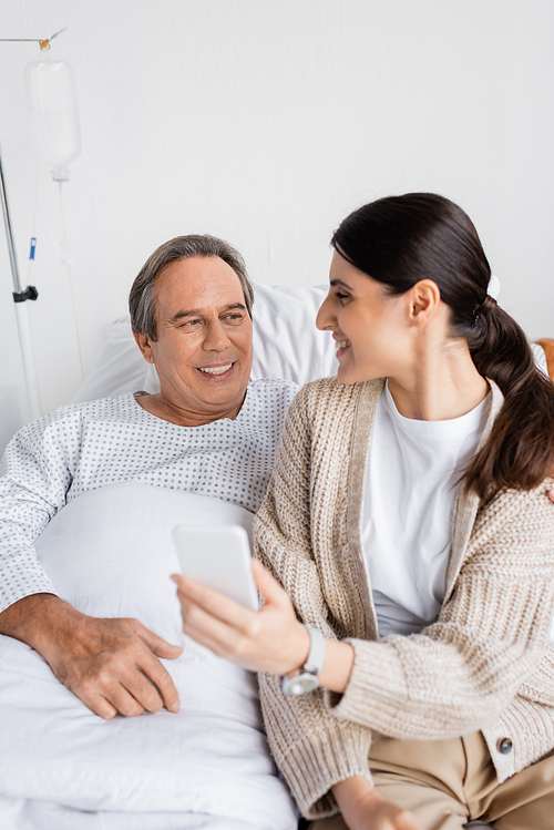 Senior patient looking at smiling daughter with smartphone in hospital ward