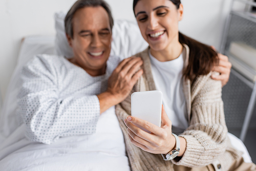 Blurred woman holding smartphone near elderly father on bed in hospital