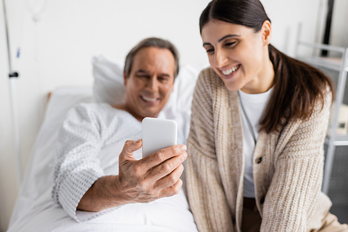 Blurred senior patient holding smartphone near daughter in hospital ward