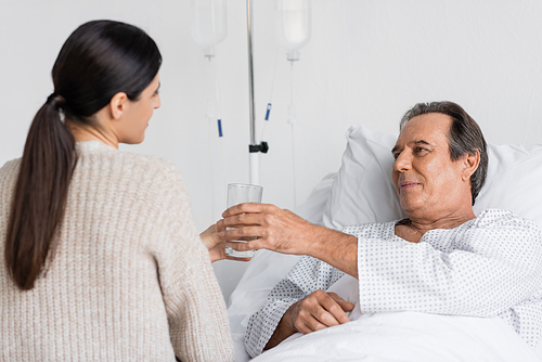 Blurred daughter giving glass of water to senior dad on hospital bed