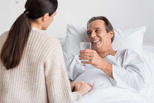 Smiling senior patient holding glass of water near blurred daughter in clinic