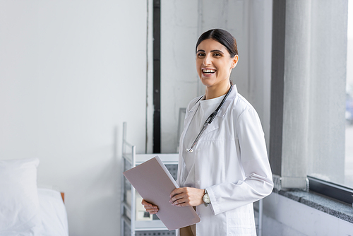 Cheerful doctor with paper folder looking at camera in hospital ward