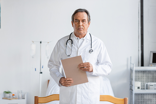 Senior doctor holding paper folder and looking at camera in clinic