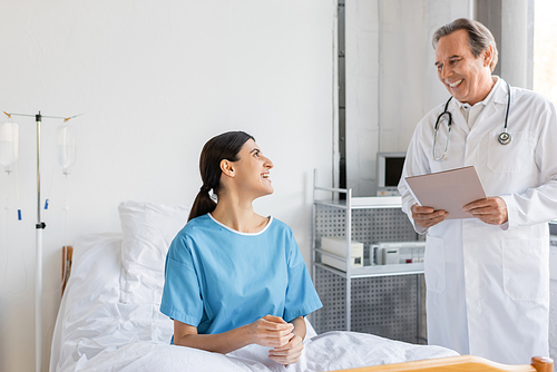 Smiling senior doctor holding paper folder near patient in hospital ward