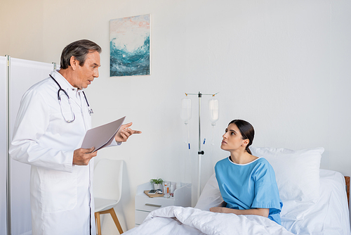 Senior doctor with paper folder talking to brunette patient in hospital ward