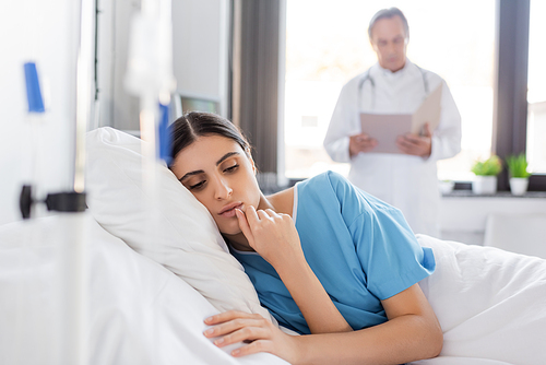 brunette woman in patient gown lying on bed near blurred doctor in clinic
