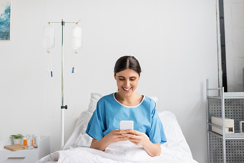 happy woman in patient gown smiling while using smartphone on bed in clinic