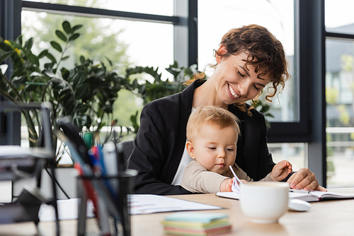 cheerful businesswoman sitting at workplace with toddler daughter near notebook and blurred coffee cup