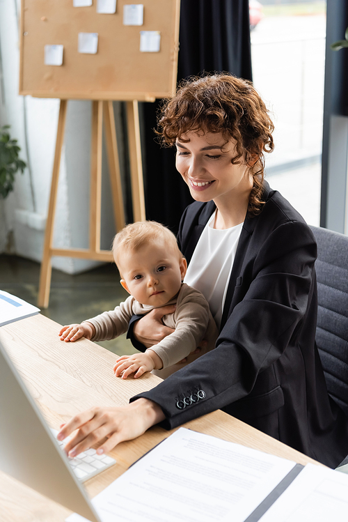 smiling businesswoman working on computer while sitting at workplace with toddler daughter