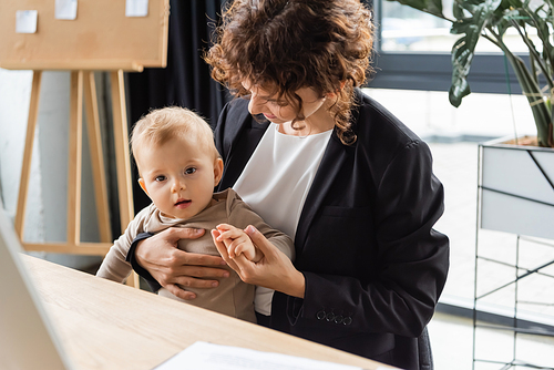 businesswoman in black blazer holding hand of toddler child while sitting at workplace in office