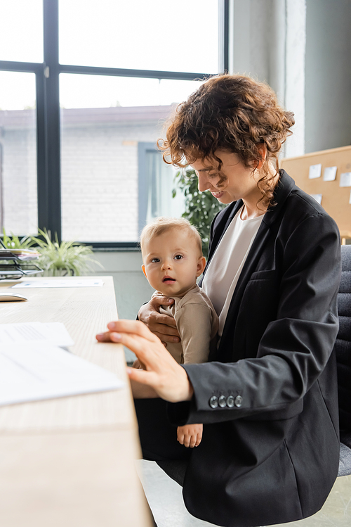 smiling businesswoman in black blazer sitting at workplace with baby while working in office