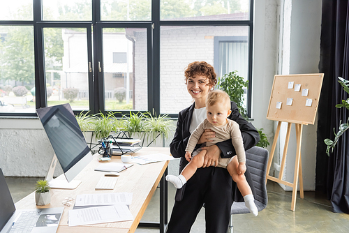 happy businesswoman standing with baby girl near work desk with computers and documents in modern office