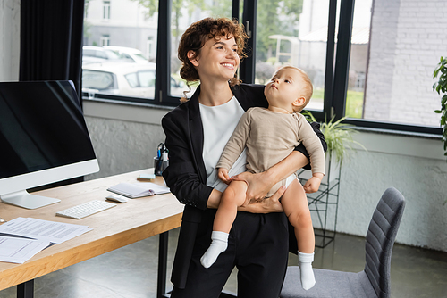 happy businesswoman looking away while standing with little child near computer monitor with blank screen