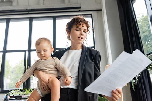 office manager in black blazer looking at papers while holding toddler baby in office