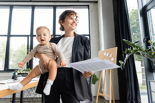happy businesswoman in black suit looking away while standing with baby and documents in office