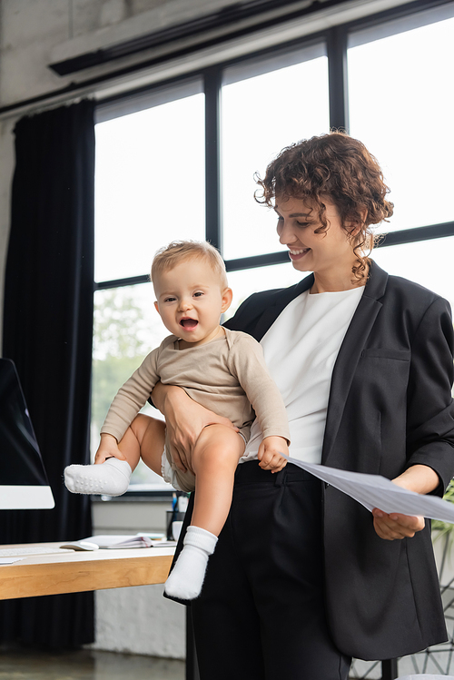 pleased businesswoman in black suit holding papers and happy daughter in romper in office