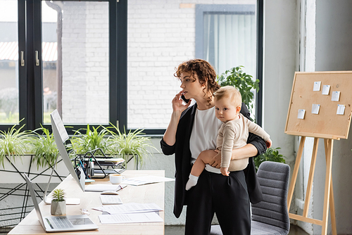 businesswoman talking on smartphone and looking at computer monitor while holding little daughter in modern office