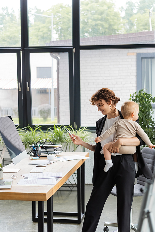 happy businesswoman standing with smartphone and baby near work desk with documents and computer monitor with blank screen