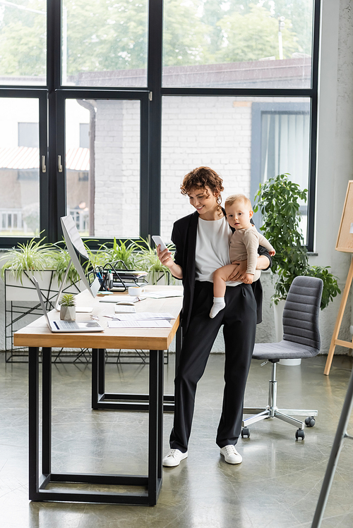 positive businesswoman holding smartphone and little child near work desk in modern office
