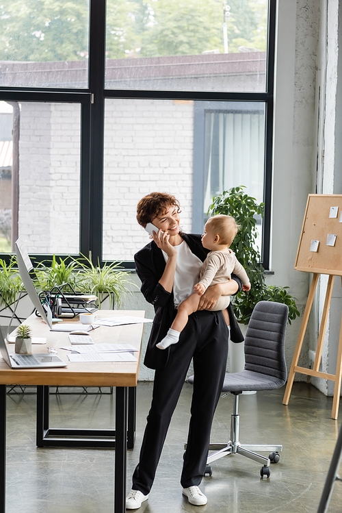 smiling businesswoman in black suit holding little daughter and talking on mobile phone in modern office