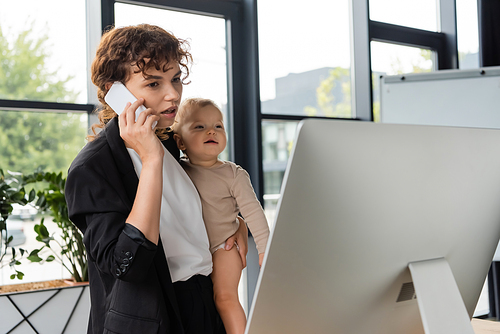 woman in black suit holding smiling baby while talking on cellphone near computer monitor