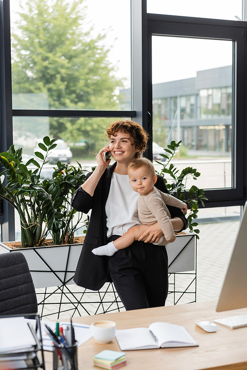 happy businesswoman looking away while holding baby and talking on mobile phone in modern office