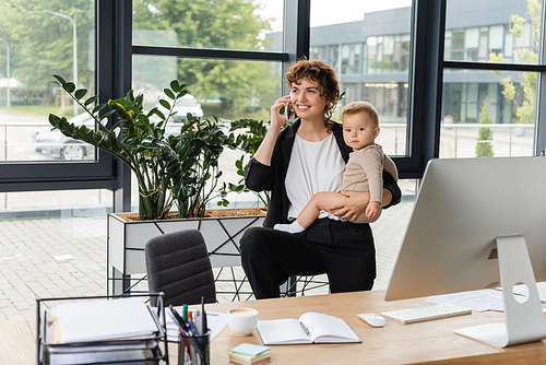 happy businesswoman holding toddler child and talking on smartphone near work desk and green plants in modern office